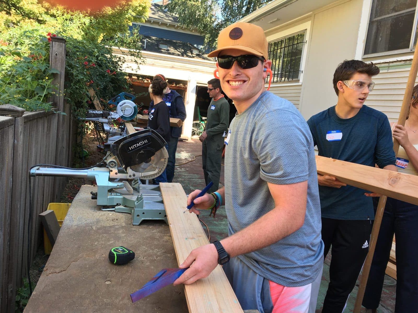 A teenaged boy wearing sunglasses and a yellow baseball cap stands next to a chopsaw. He is holding a speed square and a construction pencil over a plank of wood, and is smiling at the camera.