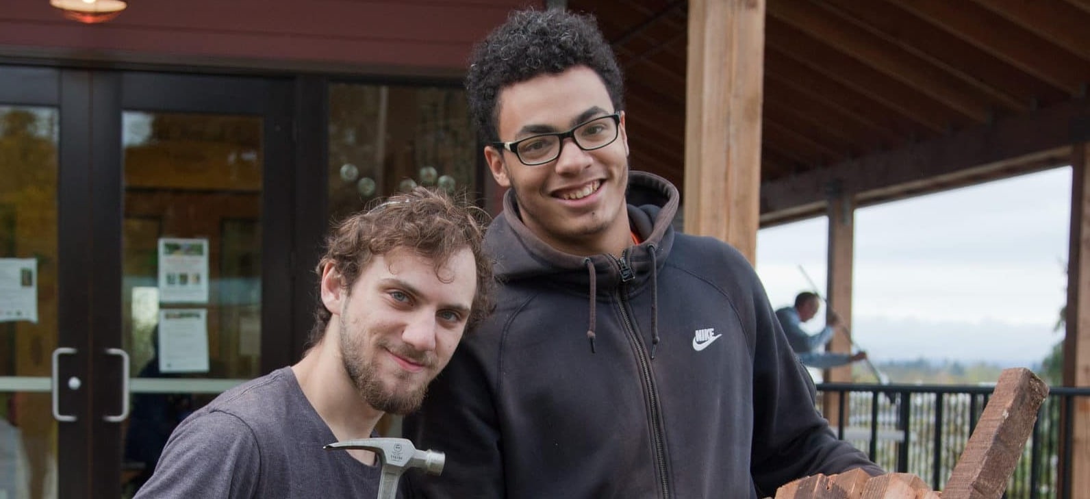 Two gap year program participants smiling at the camera, one holding a hammer and one holding wood